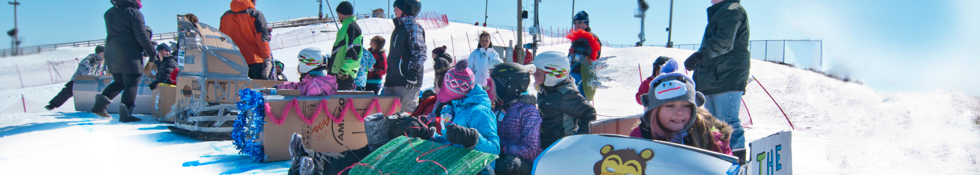 Several cardboard toboggans with kids in them, all are lined up at the start line of the race